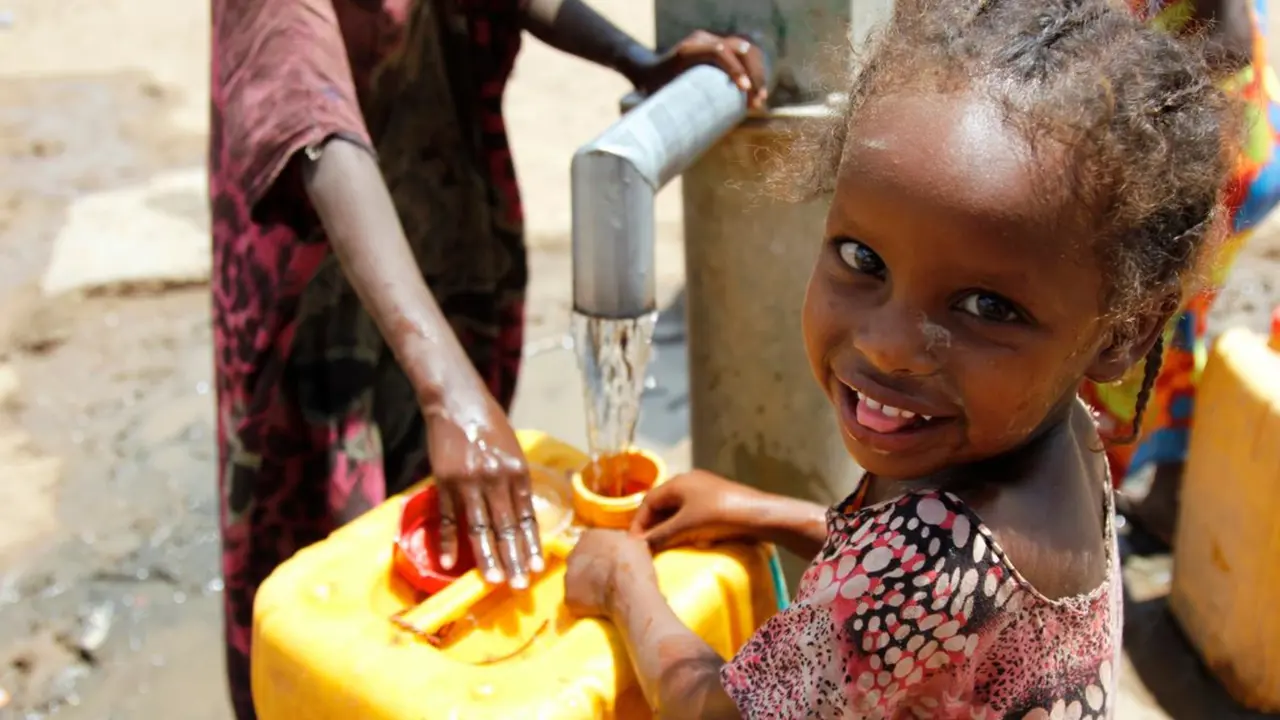 A young child looking at the camera while filling a yellow water container from a tap