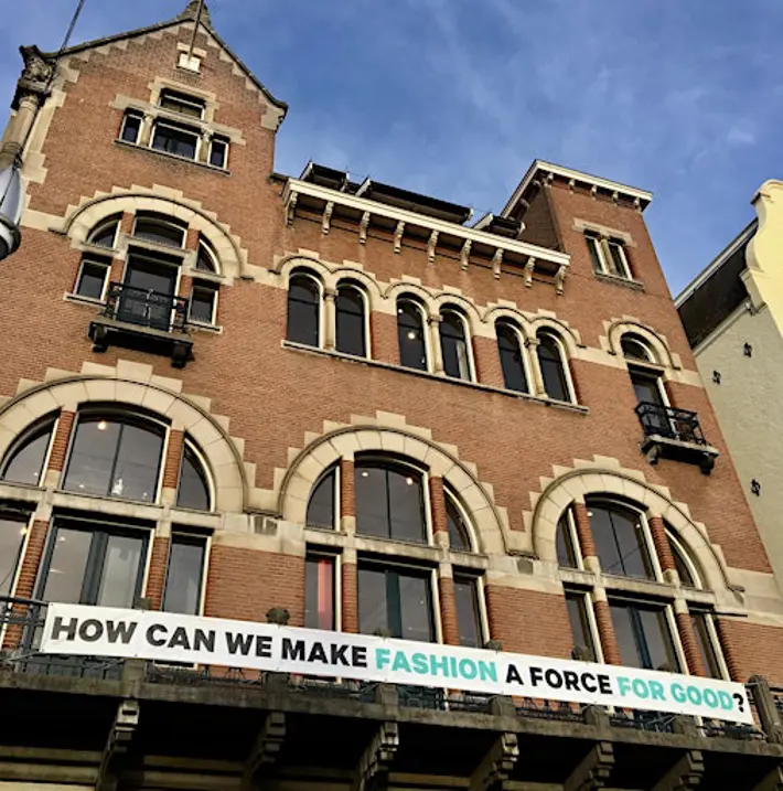 The facade of a brick building with decorative window arches and a banner that reads 'how can we make fashion a force for good?'