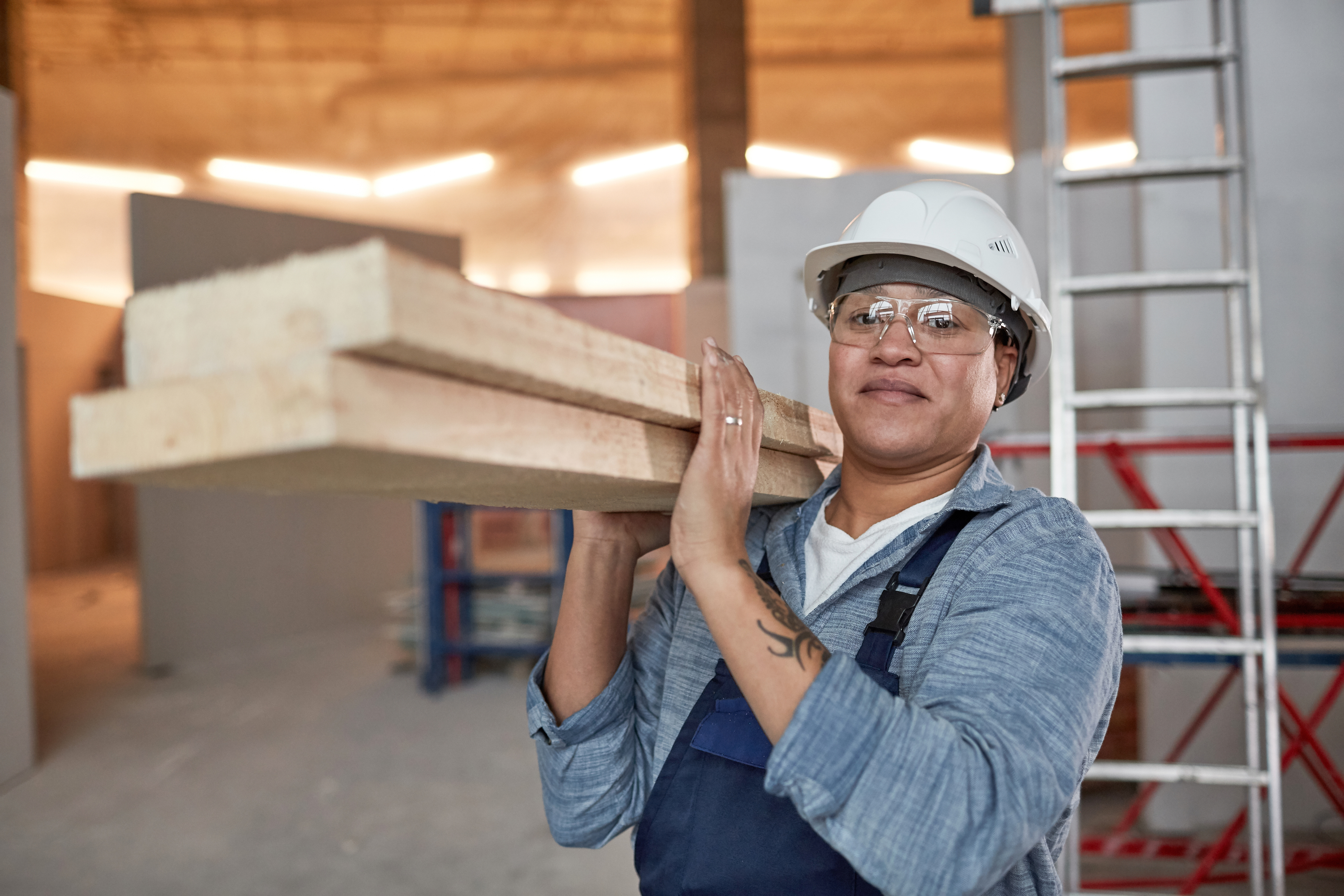 A person in a hard hat and safety glasses carrying two planks of wood over their shoulder