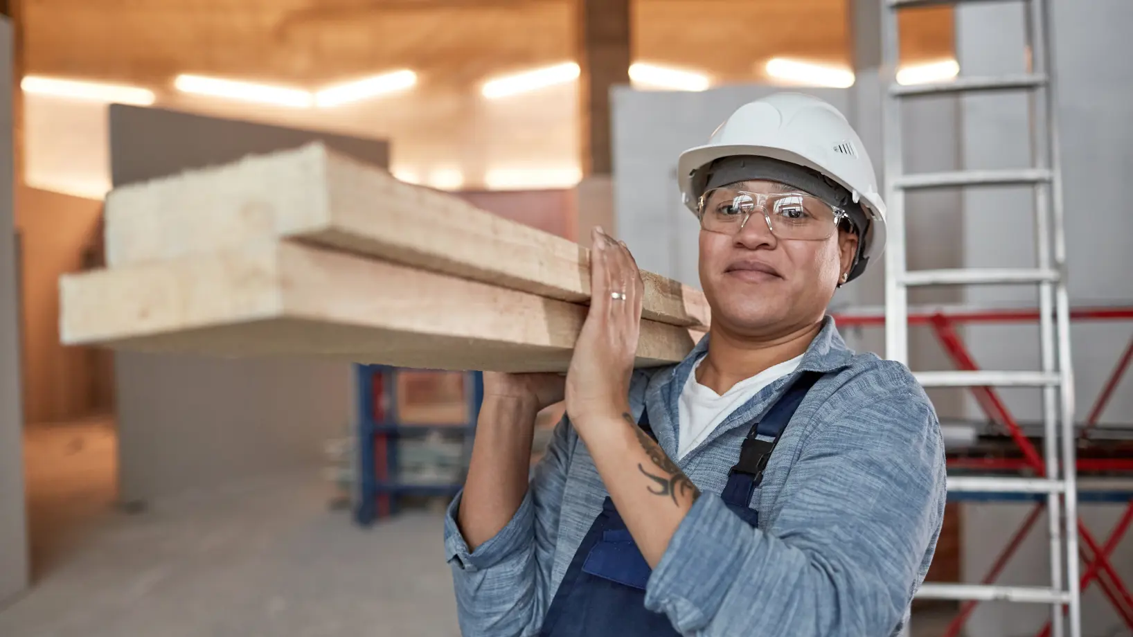 A person in a hard hat and safety glasses carrying two planks of wood over their shoulder