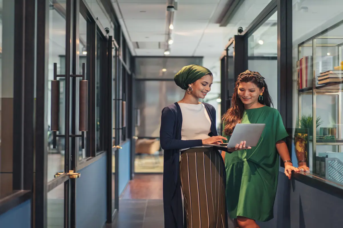 Two women standing in the corridor of an office building smiling and looking at a laptop screen.