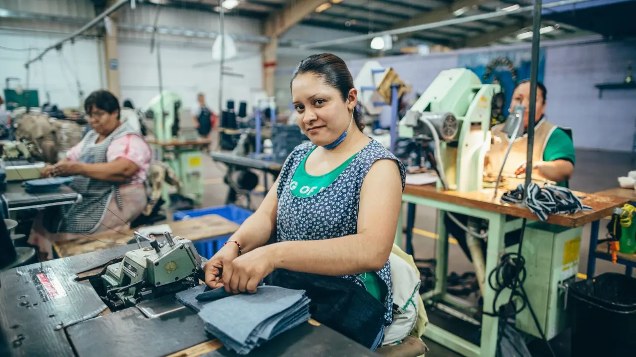 A woman looking at the camera arranging a pile of materials at a work station in a textile factory