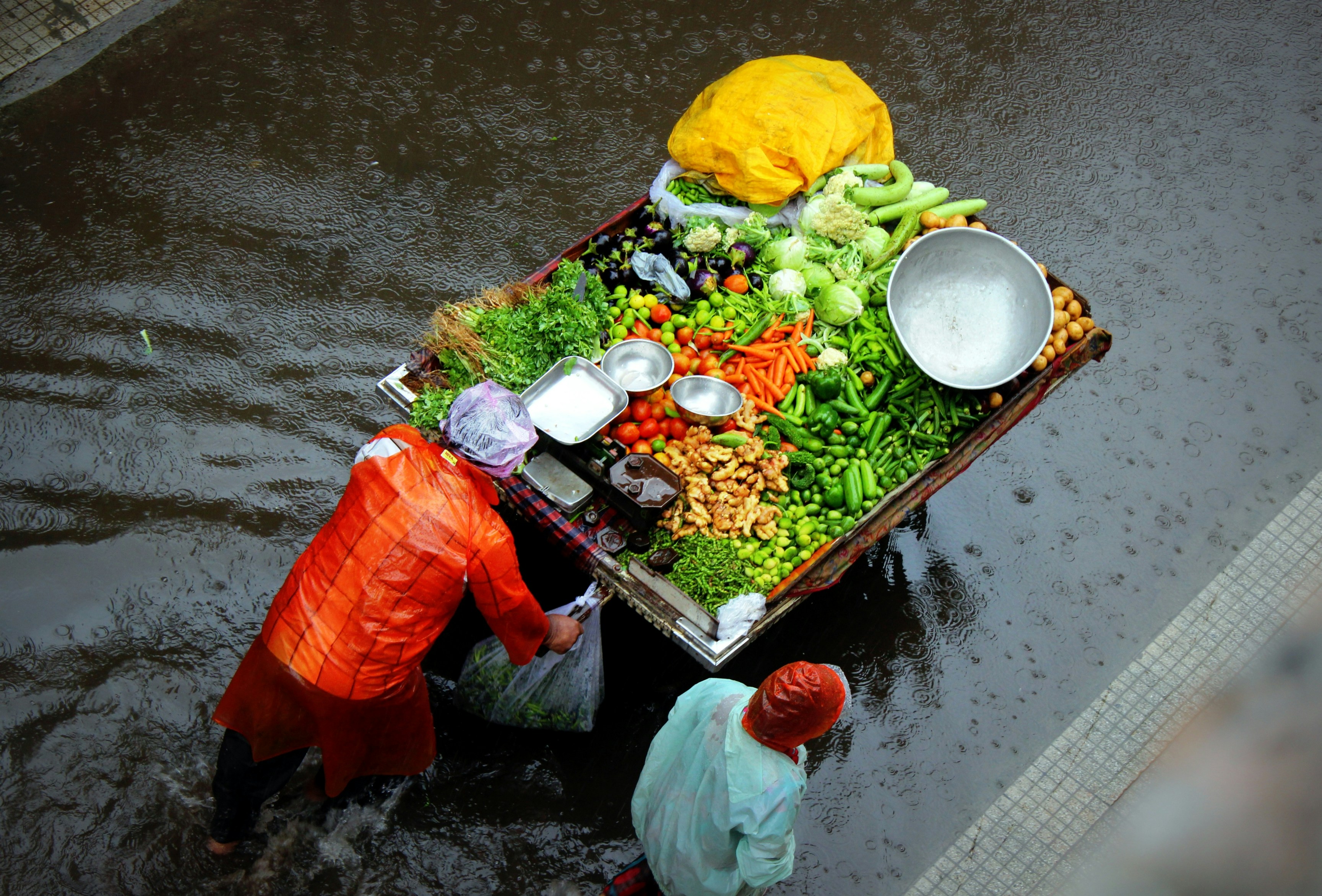 An aerial view of people pushing a card full of fresh and brightly coloured fruit and vegetables through a rainy and slightly flooded street