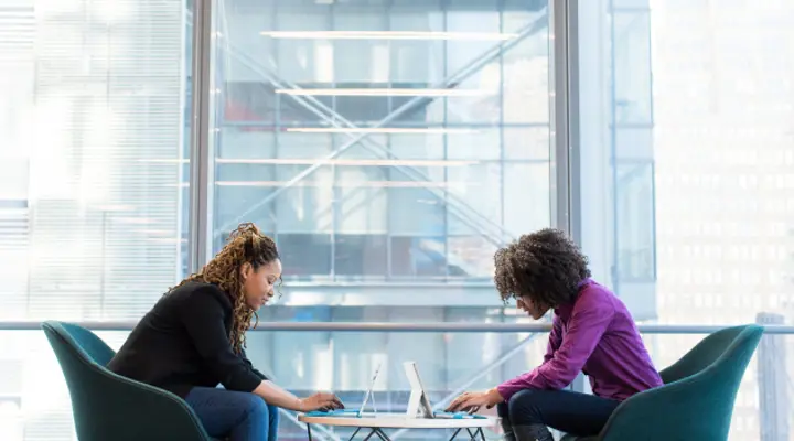 Two women at a table using tablet devices in front of a large window wall looking out onto a neighbouring high rise glass building