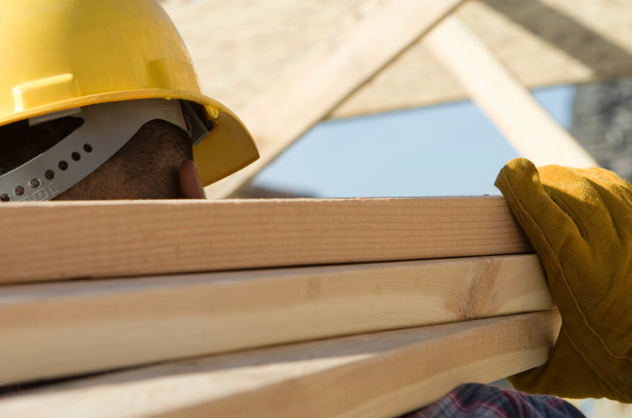 A closeup from over the shoulder of a person carrying planks of wood, they are wearing a yellow hard hat and safety gloves.