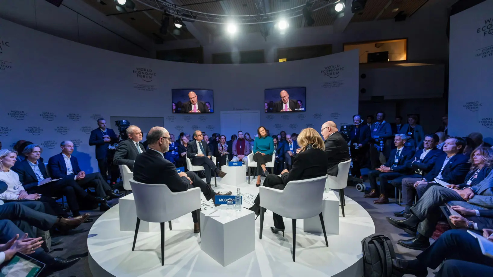 Six people on chairs having a discussion on a circular stage in a studio, with an audience surrounding them